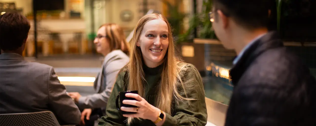 Two colleagues together shown from the males perspective, having a conversation in a plant-filled office, with blonde female colleague featured and smiling.