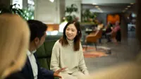 Three colleagues sitting together on dark green sofas having a conversation in a plant-filled office. Main focus is woman with short brown hair in neutral tone clothing.