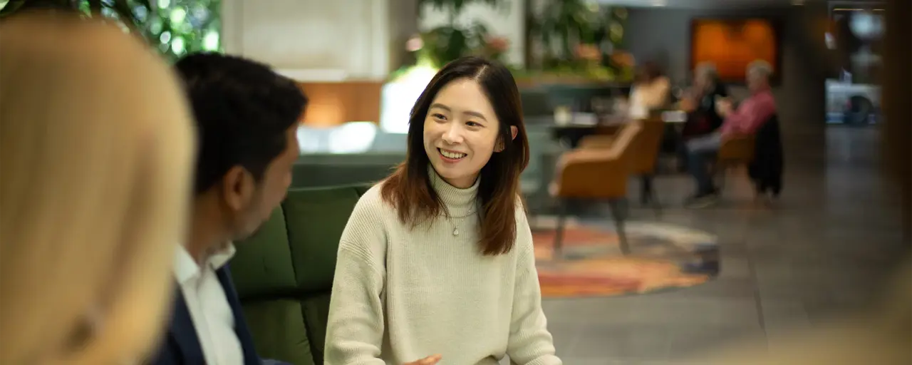 Three colleagues sitting together on dark green sofas having a conversation in a plant-filled office. Main focus is woman with short brown hair in neutral tone clothing.