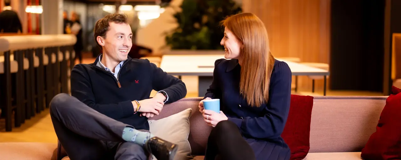 Male and female colleagues sitting together on light pink sofas in break out room having a casual chat.